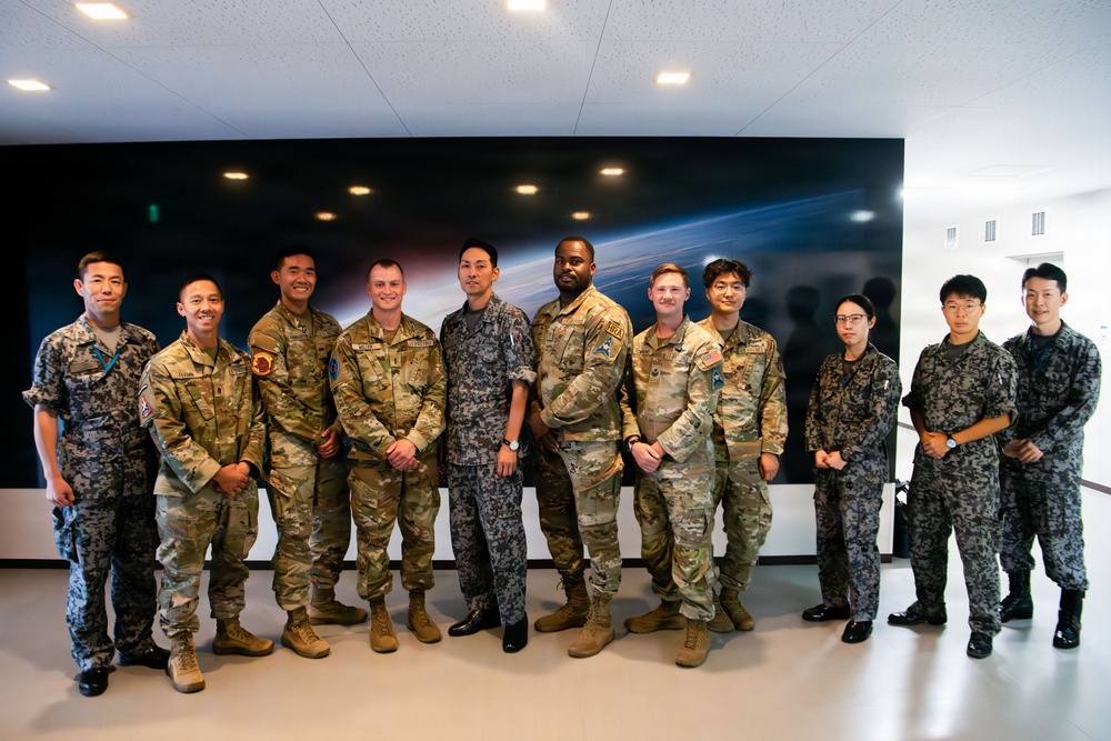 A group of approximately eleven military personnel pose together for a formal group photograph in what appears to be a modern facility. The group includes members wearing at least two distinct camouflage uniform patterns, suggesting a multinational or joint military gathering. A large space-themed display screen is visible on the wall behind them.