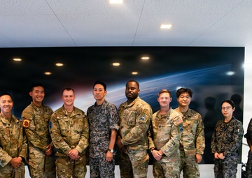 A group of approximately eleven military personnel pose together for a formal group photograph in what appears to be a modern facility. The group includes members wearing at least two distinct camouflage uniform patterns, suggesting a multinational or joint military gathering. A large space-themed display screen is visible on the wall behind them.