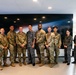 A group of approximately eleven military personnel pose together for a formal group photograph in what appears to be a modern facility. The group includes members wearing at least two distinct camouflage uniform patterns, suggesting a multinational or joint military gathering. A large space-themed display screen is visible on the wall behind them.