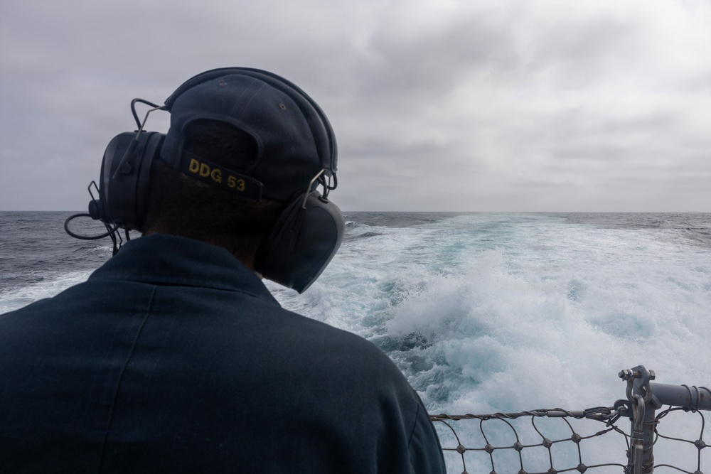 A naval personnel member is seen from behind standing on the stern of a U.S. Navy vessel, wearing hearing protection headset and a cap marked 'DDG 53'. The ship is underway at sea, leaving a prominent white wake behind it. The weather appears overcast with grey skies and choppy ocean conditions.