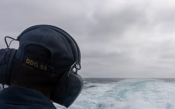 A naval personnel member is seen from behind standing on the stern of a U.S. Navy vessel, wearing hearing protection headset and a cap marked 'DDG 53'. The ship is underway at sea, leaving a prominent white wake behind it. The weather appears overcast with grey skies and choppy ocean conditions.