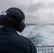 A naval personnel member is seen from behind standing on the stern of a U.S. Navy vessel, wearing hearing protection headset and a cap marked 'DDG 53'. The ship is underway at sea, leaving a prominent white wake behind it. The weather appears overcast with grey skies and choppy ocean conditions.