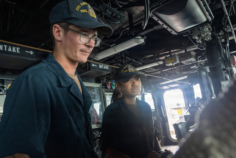 Two U.S. Navy sailors are standing on the bridge or interior of a naval vessel, both wearing matching dark navy caps with 'USS JOHN' and 'DDG' insignia visible. The male sailor on the left wears glasses and a dark blue uniform, while the female sailor stands slightly behind him. They appear to be on duty, observing or monitoring something off-camera.