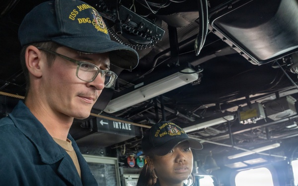 Two U.S. Navy sailors are standing on the bridge or interior of a naval vessel, both wearing matching dark navy caps with 'USS JOHN' and 'DDG' insignia visible. The male sailor on the left wears glasses and a dark blue uniform, while the female sailor stands slightly behind him. They appear to be on duty, observing or monitoring something off-camera.