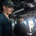 Two U.S. Navy sailors are standing on the bridge or interior of a naval vessel, both wearing matching dark navy caps with 'USS JOHN' and 'DDG' insignia visible. The male sailor on the left wears glasses and a dark blue uniform, while the female sailor stands slightly behind him. They appear to be on duty, observing or monitoring something off-camera.