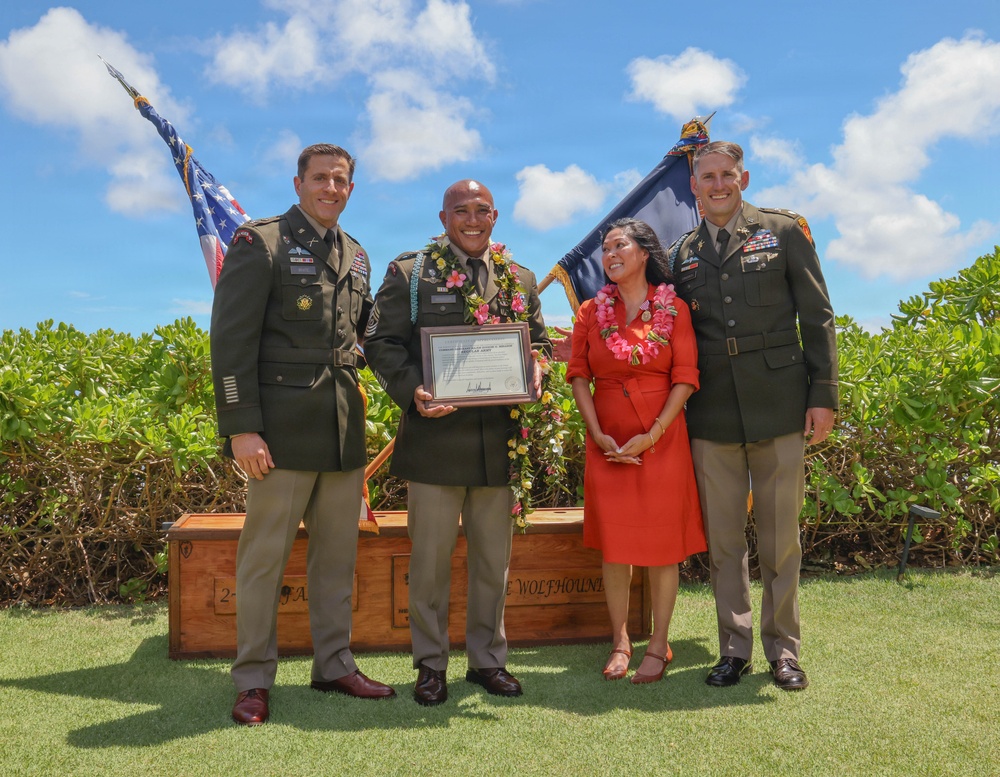 A military ceremony photographed outdoors in what appears to be Hawaii, featuring four individuals posing together. A soldier in the center is holding a framed certificate or award document while wearing traditional Hawaiian leis. Two uniformed U.S. Army soldiers flank the honoree and a woman in a red dress, with an American flag and a unit flag visible in the background.
