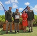 A military ceremony photographed outdoors in what appears to be Hawaii, featuring four individuals posing together. A soldier in the center is holding a framed certificate or award document while wearing traditional Hawaiian leis. Two uniformed U.S. Army soldiers flank the honoree and a woman in a red dress, with an American flag and a unit flag visible in the background.