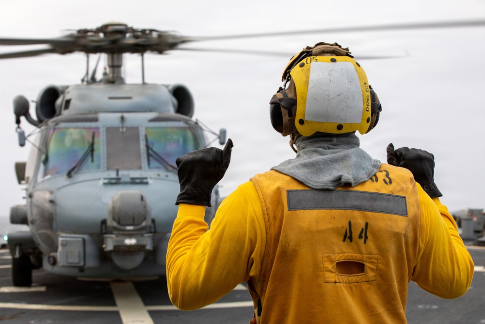 A naval deck crew member in yellow safety gear is directing a military helicopter on what appears to be a ship's flight deck. The handler is using hand signals to guide the aircraft, which has its rotors spinning. The overcast sky and ship deck environment indicate an active naval flight operations scenario.