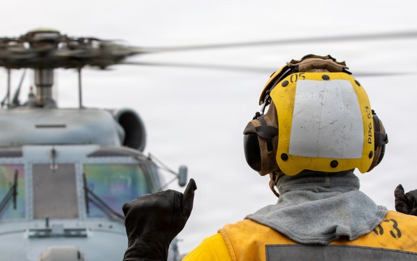 A naval deck crew member in yellow safety gear is directing a military helicopter on what appears to be a ship's flight deck. The handler is using hand signals to guide the aircraft, which has its rotors spinning. The overcast sky and ship deck environment indicate an active naval flight operations scenario.