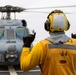 A naval deck crew member in yellow safety gear is directing a military helicopter on what appears to be a ship's flight deck. The handler is using hand signals to guide the aircraft, which has its rotors spinning. The overcast sky and ship deck environment indicate an active naval flight operations scenario.