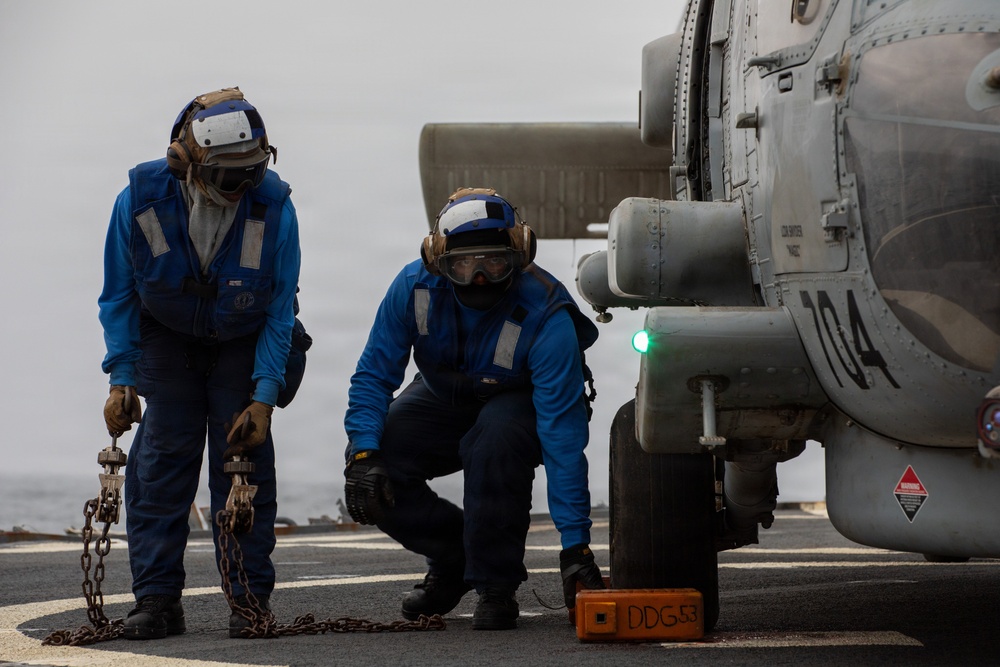 Two naval personnel in blue flight deck coveralls are working on a helicopter aboard a U.S. Navy ship. One sailor holds tie-down chains while the other crouches near a wheel chock marked 'DDG 53', indicating operations aboard USS John Paul Jones or a similarly designated Arleigh Burke-class destroyer. The helicopter, numbered '104', has a green navigation light illuminated on its fuselage.