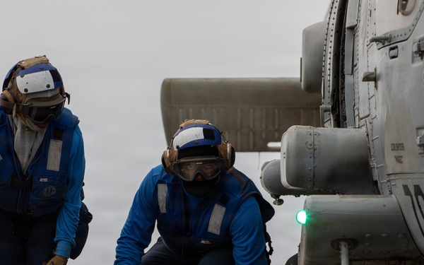 Two naval personnel in blue flight deck coveralls are working on a helicopter aboard a U.S. Navy ship. One sailor holds tie-down chains while the other crouches near a wheel chock marked 'DDG 53', indicating operations aboard USS John Paul Jones or a similarly designated Arleigh Burke-class destroyer. The helicopter, numbered '104', has a green navigation light illuminated on its fuselage.