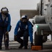 Two naval personnel in blue flight deck coveralls are working on a helicopter aboard a U.S. Navy ship. One sailor holds tie-down chains while the other crouches near a wheel chock marked 'DDG 53', indicating operations aboard USS John Paul Jones or a similarly designated Arleigh Burke-class destroyer. The helicopter, numbered '104', has a green navigation light illuminated on its fuselage.