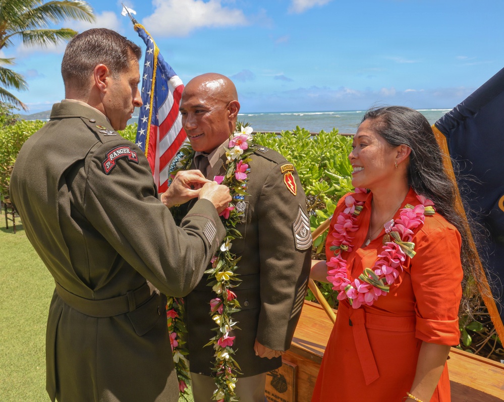 A military ceremony taking place outdoors in what appears to be Hawaii, where a senior officer is pinning a medal or award onto a non-commissioned officer wearing traditional Hawaiian leis. A woman in an orange dress, also wearing a lei, stands nearby smiling. The ocean and tropical vegetation are visible in the background.