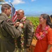 A military ceremony taking place outdoors in what appears to be Hawaii, where a senior officer is pinning a medal or award onto a non-commissioned officer wearing traditional Hawaiian leis. A woman in an orange dress, also wearing a lei, stands nearby smiling. The ocean and tropical vegetation are visible in the background.