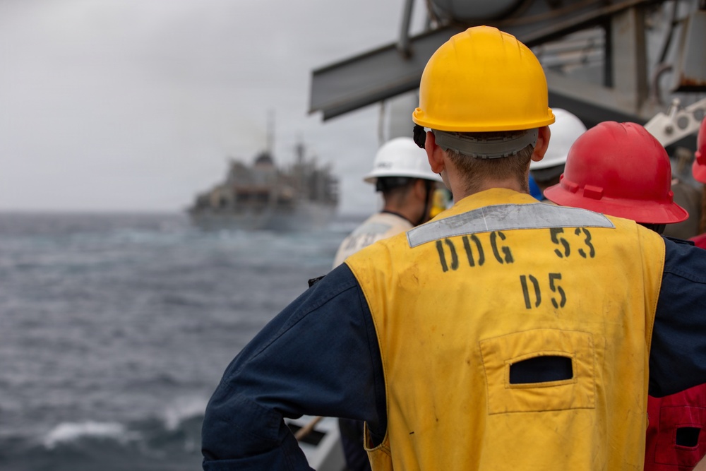 Naval personnel aboard USS John Paul Jones (DDG-53) observe a replenishment-at-sea (RAS) operation with another vessel visible in the background. Crew members are wearing color-coded safety helmets and high-visibility vests, standard protocol during underway replenishment operations. The scene takes place on open ocean under overcast conditions.