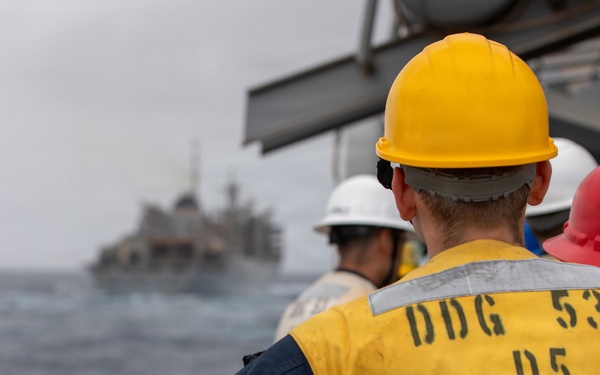 Naval personnel aboard USS John Paul Jones (DDG-53) observe a replenishment-at-sea (RAS) operation with another vessel visible in the background. Crew members are wearing color-coded safety helmets and high-visibility vests, standard protocol during underway replenishment operations. The scene takes place on open ocean under overcast conditions.