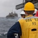 Naval personnel aboard USS John Paul Jones (DDG-53) observe a replenishment-at-sea (RAS) operation with another vessel visible in the background. Crew members are wearing color-coded safety helmets and high-visibility vests, standard protocol during underway replenishment operations. The scene takes place on open ocean under overcast conditions.
