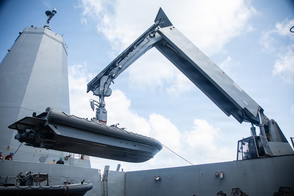 A rigid inflatable boat (RIB) is being lifted or lowered by a crane davit system aboard a naval vessel. The image is taken from a low angle looking upward, showing the ship's superstructure, crane arm, and the suspended boat against a partly cloudy sky. At least two crew members are visible on deck during the operation.