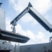 A rigid inflatable boat (RIB) is being lifted or lowered by a crane davit system aboard a naval vessel. The image is taken from a low angle looking upward, showing the ship's superstructure, crane arm, and the suspended boat against a partly cloudy sky. At least two crew members are visible on deck during the operation.