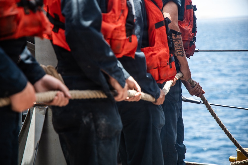 A line of personnel wearing orange life vests and dark uniforms are standing on the deck of a vessel, collectively pulling a thick rope. The scene appears to be a maritime operation, likely a line-handling or mooring exercise. One individual on the right has a visible tattoo on their forearm in a gothic/script style.