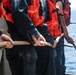 A line of personnel wearing orange life vests and dark uniforms are standing on the deck of a vessel, collectively pulling a thick rope. The scene appears to be a maritime operation, likely a line-handling or mooring exercise. One individual on the right has a visible tattoo on their forearm in a gothic/script style.