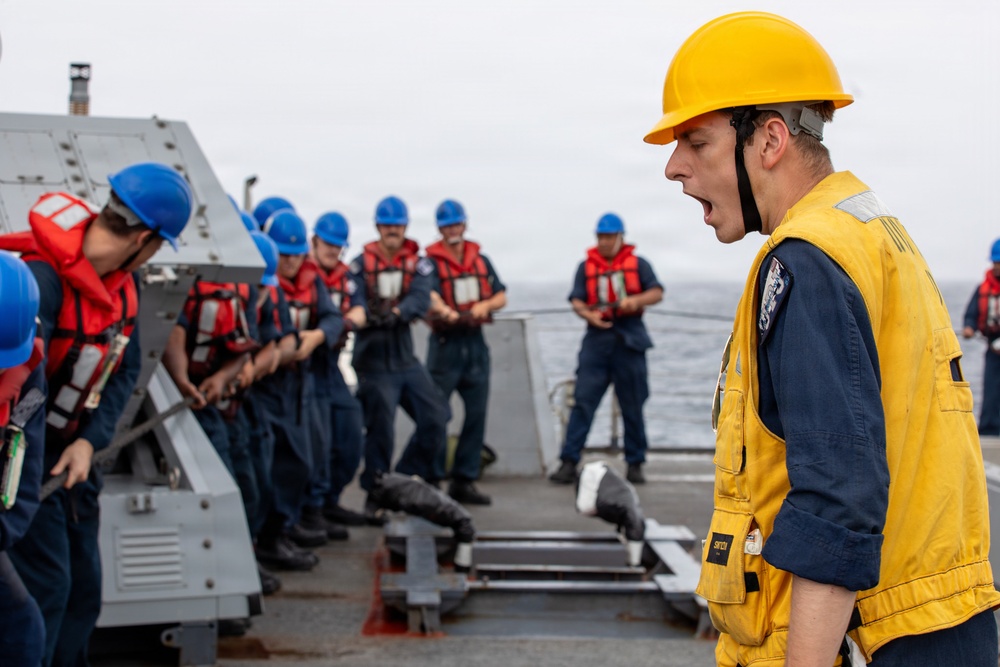 A naval officer or supervisor wearing a yellow hard hat and yellow safety vest is shouting commands to a group of sailors on the deck of a naval vessel. The sailors are wearing blue uniforms, red life vests, and blue hard hats, and appear to be engaged in a deck operation or drill. The scene takes place at sea, with ocean visible in the background.