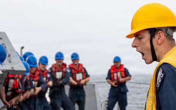 A naval officer or supervisor wearing a yellow hard hat and yellow safety vest is shouting commands to a group of sailors on the deck of a naval vessel. The sailors are wearing blue uniforms, red life vests, and blue hard hats, and appear to be engaged in a deck operation or drill. The scene takes place at sea, with ocean visible in the background.