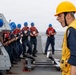A naval officer or supervisor wearing a yellow hard hat and yellow safety vest is shouting commands to a group of sailors on the deck of a naval vessel. The sailors are wearing blue uniforms, red life vests, and blue hard hats, and appear to be engaged in a deck operation or drill. The scene takes place at sea, with ocean visible in the background.