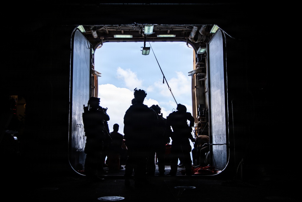 The image shows silhouetted figures in full tactical gear standing at the open rear ramp or bay door of a large military vessel or aircraft, backlit by a bright sky with clouds. The subjects are rendered almost entirely in silhouette due to strong backlighting, creating a high-contrast composition. The setting appears to be a military operation or exercise involving personnel prepared for deployment.