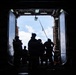 The image shows silhouetted figures in full tactical gear standing at the open rear ramp or bay door of a large military vessel or aircraft, backlit by a bright sky with clouds. The subjects are rendered almost entirely in silhouette due to strong backlighting, creating a high-contrast composition. The setting appears to be a military operation or exercise involving personnel prepared for deployment.