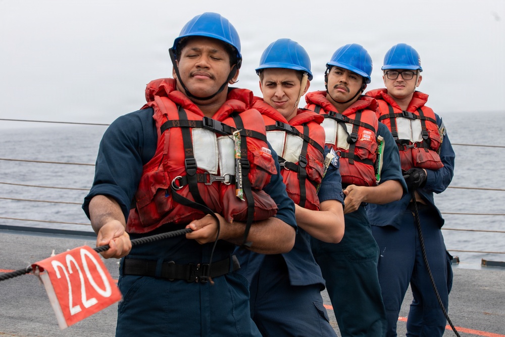 Four naval personnel in blue coveralls, red life vests, and blue hard hats are pulling a rope on the deck of a ship at sea. The lead sailor is gripping a rope marked with a red pennant displaying the number '220'. The team appears to be engaged in a line-handling or mooring exercise aboard a military vessel.