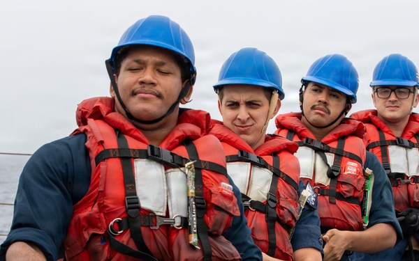 Four naval personnel in blue coveralls, red life vests, and blue hard hats are pulling a rope on the deck of a ship at sea. The lead sailor is gripping a rope marked with a red pennant displaying the number '220'. The team appears to be engaged in a line-handling or mooring exercise aboard a military vessel.