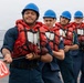 Four naval personnel in blue coveralls, red life vests, and blue hard hats are pulling a rope on the deck of a ship at sea. The lead sailor is gripping a rope marked with a red pennant displaying the number '220'. The team appears to be engaged in a line-handling or mooring exercise aboard a military vessel.