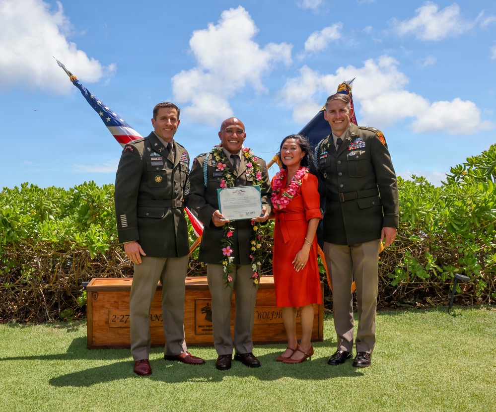 A military ceremony photograph showing four individuals posing outdoors, likely in Hawaii based on the traditional lei garlands worn. Two individuals in U.S. Army dress uniforms flank a central male soldier wearing multiple leis and holding a certificate, accompanied by a woman in a red dress also wearing a lei. A wooden crate marked '2-27' and 'WOLFHOUND' is visible in the background along with U.S. and unit flags.