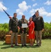 A military ceremony photograph showing four individuals posing outdoors, likely in Hawaii based on the traditional lei garlands worn. Two individuals in U.S. Army dress uniforms flank a central male soldier wearing multiple leis and holding a certificate, accompanied by a woman in a red dress also wearing a lei. A wooden crate marked '2-27' and 'WOLFHOUND' is visible in the background along with U.S. and unit flags.