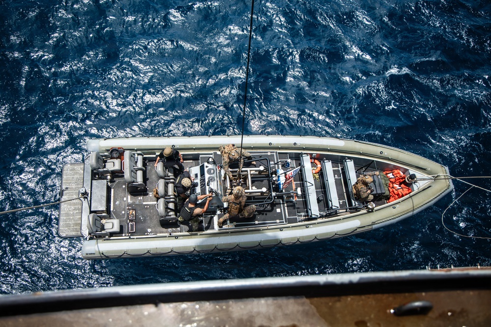 An aerial top-down view of a military rigid-hull inflatable boat (RHIB) on open ocean water. Several armed personnel in tactical gear and body armor are positioned throughout the vessel. The boat appears to be tethered to a larger vessel visible at the bottom edge of the frame.
