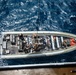 An aerial top-down view of a military rigid-hull inflatable boat (RHIB) on open ocean water. Several armed personnel in tactical gear and body armor are positioned throughout the vessel. The boat appears to be tethered to a larger vessel visible at the bottom edge of the frame.