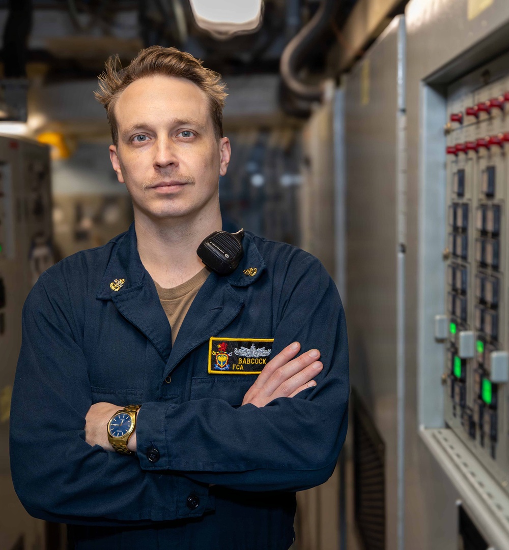 A U.S. Navy sailor identified by name tape as 'BABCOCK' with the rating designation 'FCA' poses with arms crossed aboard what appears to be a naval vessel. He is wearing a navy blue utility uniform (NWU Type III or similar) with a radio/speaker mic clipped to his shoulder and a gold wristwatch. He is standing next to what appears to be an electrical or control panel in a shipboard compartment.