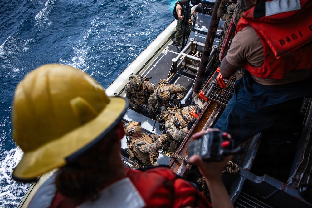 The image shows a military maritime operation where personnel in camouflage combat gear are boarding or disembarking from a small rigid-hull inflatable boat (RHIB) alongside a larger vessel. Crew members wearing red life vests and safety helmets are overseeing the transfer operation from the deck above. The scene appears to be a training exercise or tactical insertion/extraction operation on open ocean waters.