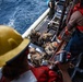 The image shows a military maritime operation where personnel in camouflage combat gear are boarding or disembarking from a small rigid-hull inflatable boat (RHIB) alongside a larger vessel. Crew members wearing red life vests and safety helmets are overseeing the transfer operation from the deck above. The scene appears to be a training exercise or tactical insertion/extraction operation on open ocean waters.