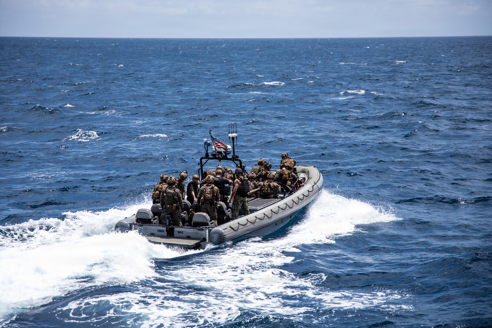 A rigid-hull inflatable boat (RHIB) carrying multiple armed military personnel in tactical gear is underway at speed on open ocean waters. The vessel displays what appears to be a US flag on a mounted pole at the center console. The personnel are equipped with tactical vests, helmets, and backpacks, consistent with a maritime special operations or boarding team exercise.