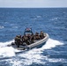 A rigid-hull inflatable boat (RHIB) carrying multiple armed military personnel in tactical gear is underway at speed on open ocean waters. The vessel displays what appears to be a US flag on a mounted pole at the center console. The personnel are equipped with tactical vests, helmets, and backpacks, consistent with a maritime special operations or boarding team exercise.