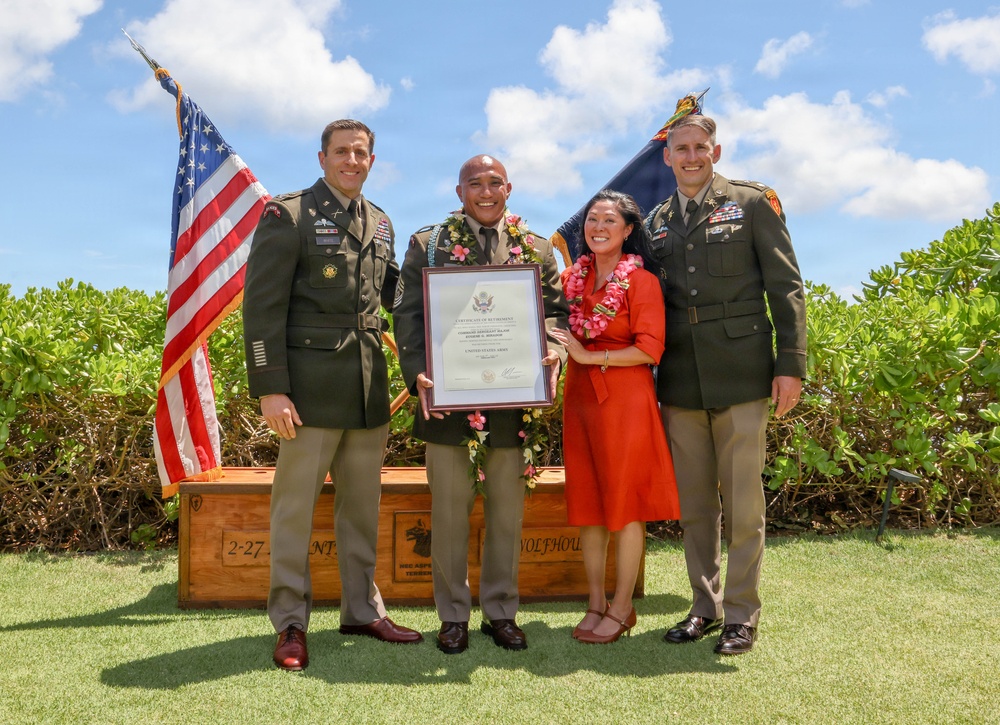 A military retirement ceremony taking place outdoors in what appears to be Hawaii, evidenced by the floral leis worn by the central figures. A service member in the center is holding a Certificate of Retirement alongside a woman in a red dress, flanked by two uniformed U.S. Army officers. An American flag and a unit guidon are visible in the background alongside a wooden crate marked '2-27' referencing a military unit.