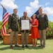 A military retirement ceremony taking place outdoors in what appears to be Hawaii, evidenced by the floral leis worn by the central figures. A service member in the center is holding a Certificate of Retirement alongside a woman in a red dress, flanked by two uniformed U.S. Army officers. An American flag and a unit guidon are visible in the background alongside a wooden crate marked '2-27' referencing a military unit.