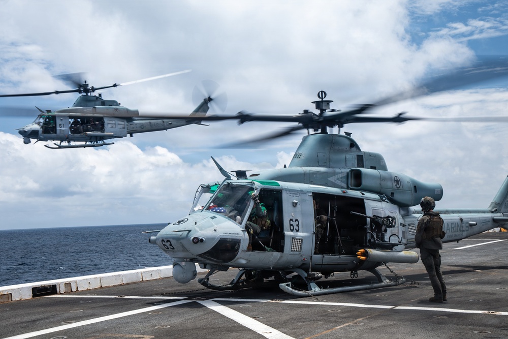 Two U.S. Marines UH-1Y Venom helicopters are operating on what appears to be the flight deck of a naval amphibious assault ship at sea. The foreground helicopter, numbered '63', is on deck with rotors spinning while a crew member stands nearby, and a second helicopter is airborne in the background. The 'MARINES' marking is visible on the fuselage, confirming U.S. Marine Corps ownership.