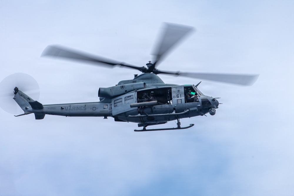 A U.S. Marines military helicopter is captured in flight against an overcast sky. The aircraft is a UH-1Y Venom (Huey) variant, identifiable by its twin-blade rotor system and 'MARINES' marking on the fuselage. The helicopter appears to be in active operation with crew members visible through the open cabin door.