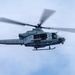 A U.S. Marines military helicopter is captured in flight against an overcast sky. The aircraft is a UH-1Y Venom (Huey) variant, identifiable by its twin-blade rotor system and 'MARINES' marking on the fuselage. The helicopter appears to be in active operation with crew members visible through the open cabin door.