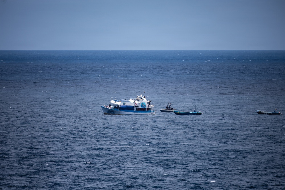 The image shows an open ocean scene with three vessels visible on deep blue water. A larger white and blue passenger or tour vessel is positioned centrally, flanked by two smaller dark-hulled rigid inflatable boats (RIBs) or patrol craft. The scene suggests a maritime operation, escort, or interception scenario.