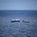 The image shows an open ocean scene with three vessels visible on deep blue water. A larger white and blue passenger or tour vessel is positioned centrally, flanked by two smaller dark-hulled rigid inflatable boats (RIBs) or patrol craft. The scene suggests a maritime operation, escort, or interception scenario.